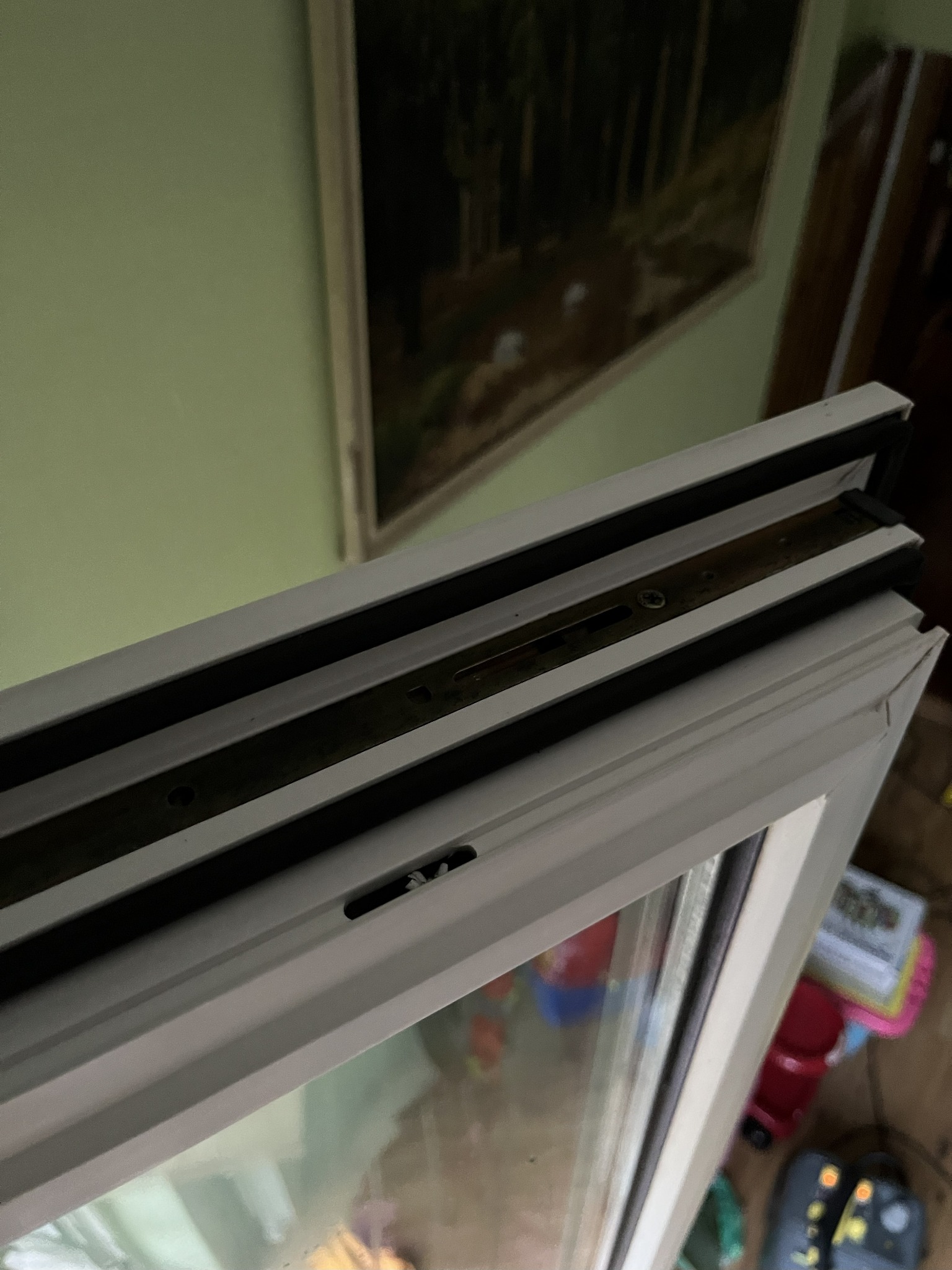 Otwarty fragment okna PCV z widocznym mechanizmem, uszczelkami i szybą, na tle ściany i obrazu. Widoczne zabawki w tle. Ujęcie z bliska, z góry.