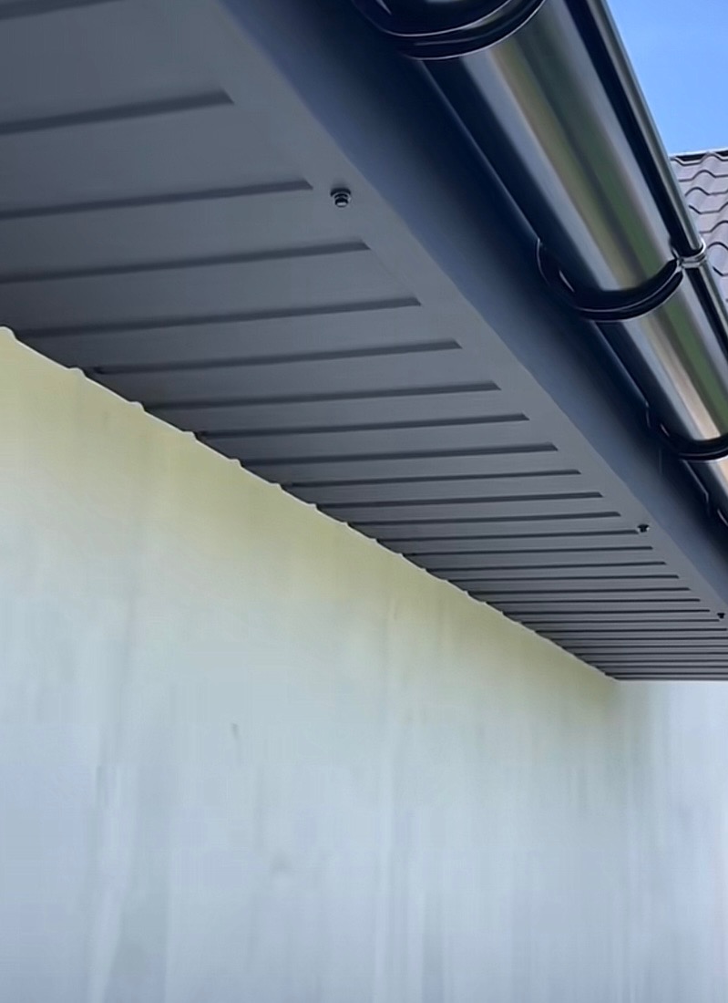 Nowoczesny, grafitowy system orynnowania zamontowany przy podbitce dachowej z widocznymi mocowaniami. Tło stanowi jasna ściana budynku i fragment dachu z dachówką.