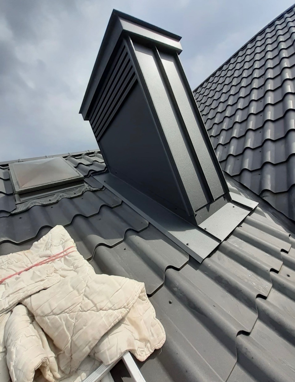 Szary dach z dachówki z obróbką blacharską komina wentylacyjnego i oknem dachowym. Na dachu leży koc. Ujęcie z dołu, pochmurne niebo w tle.