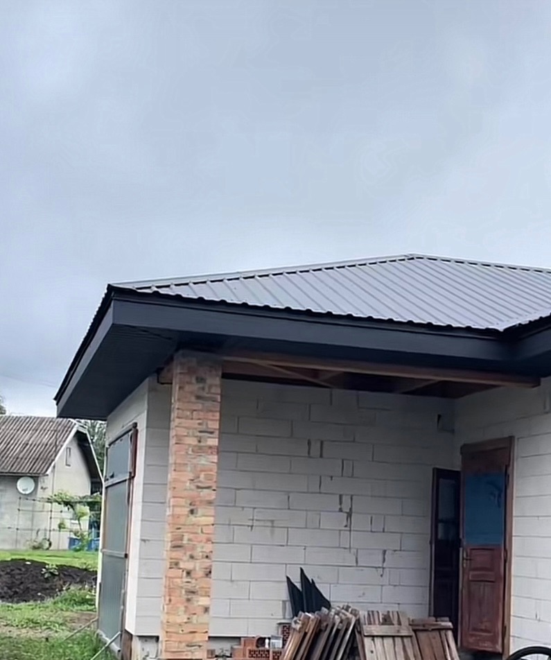 Szary dach z blachy trapezowej i czarne rynny na budynku z białego bloczka, widok z dołu, pochmurne niebo w tle.