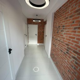 AM Projekt Usługi remontowo- wykończeniowe - Korytarz po remoncie: białe ściany, błyszcząca podłoga z płytek, ściana z cegły, okrągłe lampy sufitowe. Widok na drzwi i dalszą część korytarza.
