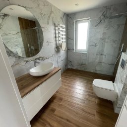 AM Projekt Usługi remontowo- wykończeniowe - Nowoczesna łazienka z marmurowymi płytkami, drewnopodobną podłogą i owalną umywalką nablatową. Widoczny grzejnik drabinkowy i kabina prysznicowa.