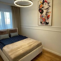 AM Projekt Usługi remontowo- wykończeniowe - Jasna sypialnia z beżowym łóżkiem, dekoracyjną sztukaterią ścienną i obrazem kobiety z kwiatami. Okrągła lampa sufitowa dodaje nowoczesnego akcentu.