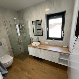 AM Projekt Usługi remontowo- wykończeniowe - Nowoczesna łazienka z kabiną prysznicową, umywalką nablatową na szafce z drewnianym blatem i płytkami imitującymi marmur. Okno z czarną ramą.