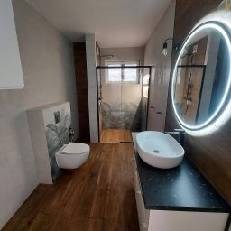 AM Projekt Usługi remontowo- wykończeniowe - Nowoczesna łazienka z drewnianą podłogą i akcentami, kabina prysznicowa z motywem roślinnym, okrągłe lustro z podświetleniem LED i wisząca toaleta.