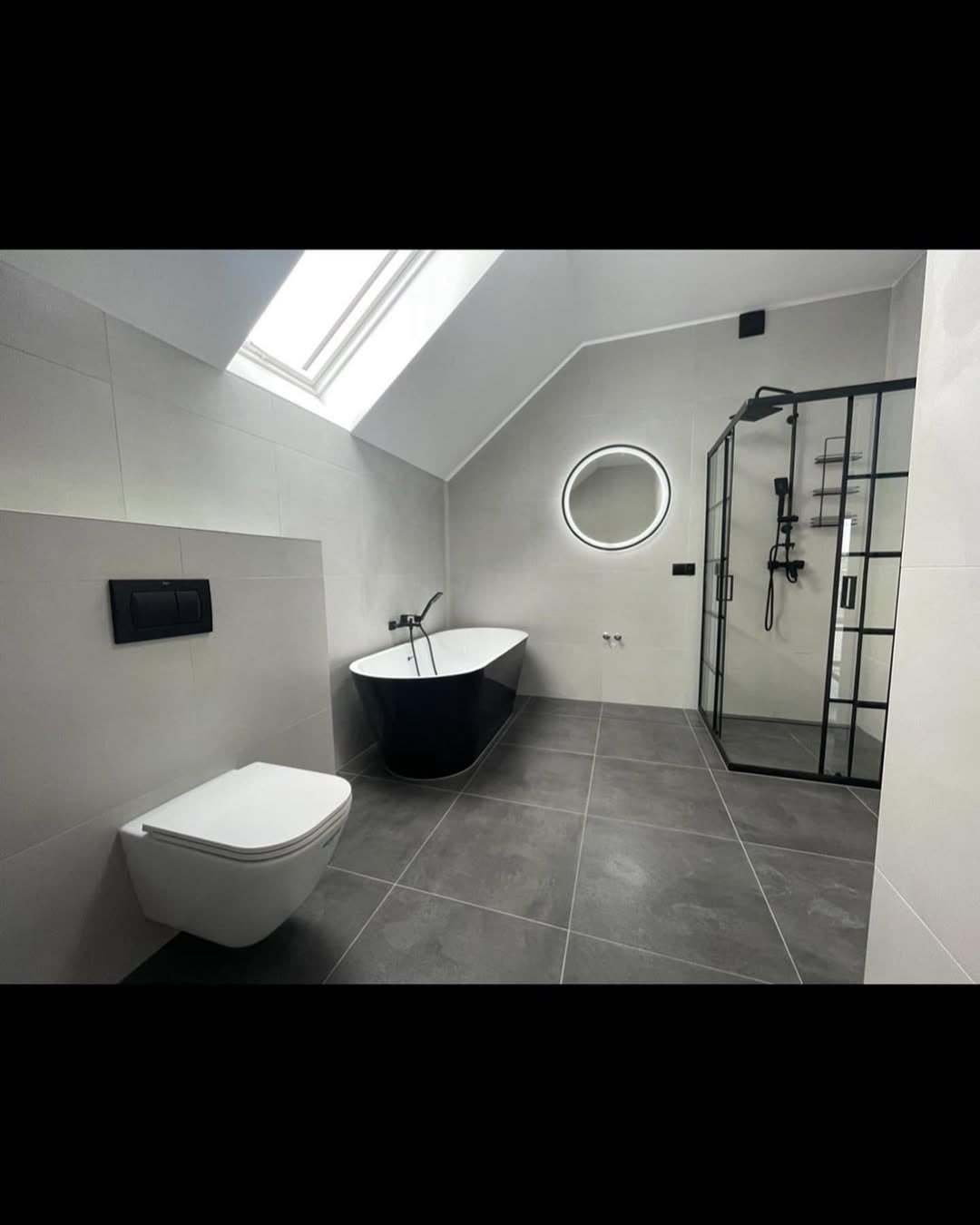 Stylowa łazienka na poddaszu z czarną wanną wolnostojącą, kabiną prysznicową z czarnymi profilami i okrągłym lustrem z podświetleniem LED.