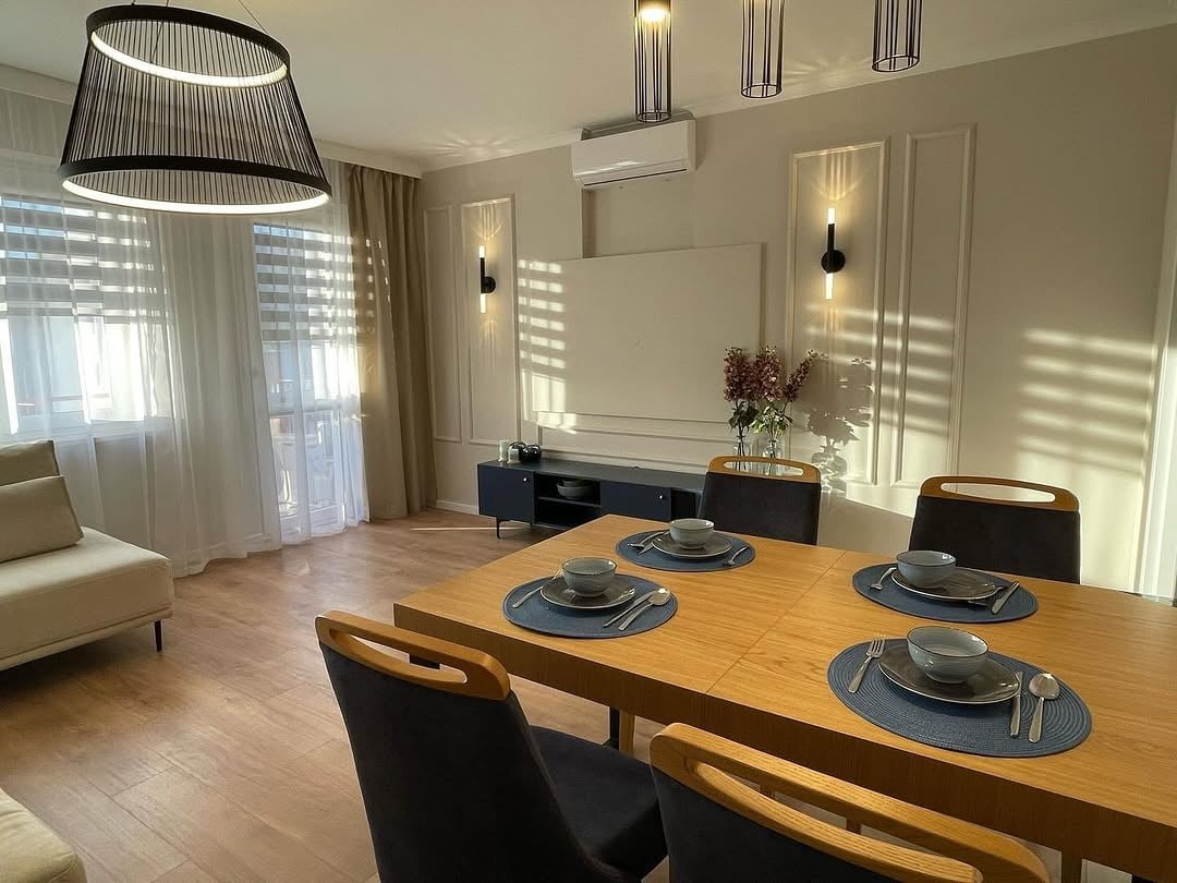 Jasna jadalnia z drewnianym stołem, nakrytym dla czterech osób. W tle okno z żaluzjami i zasłonami, designerskie lampy i dekoracyjne panele ścienne z oświetleniem.