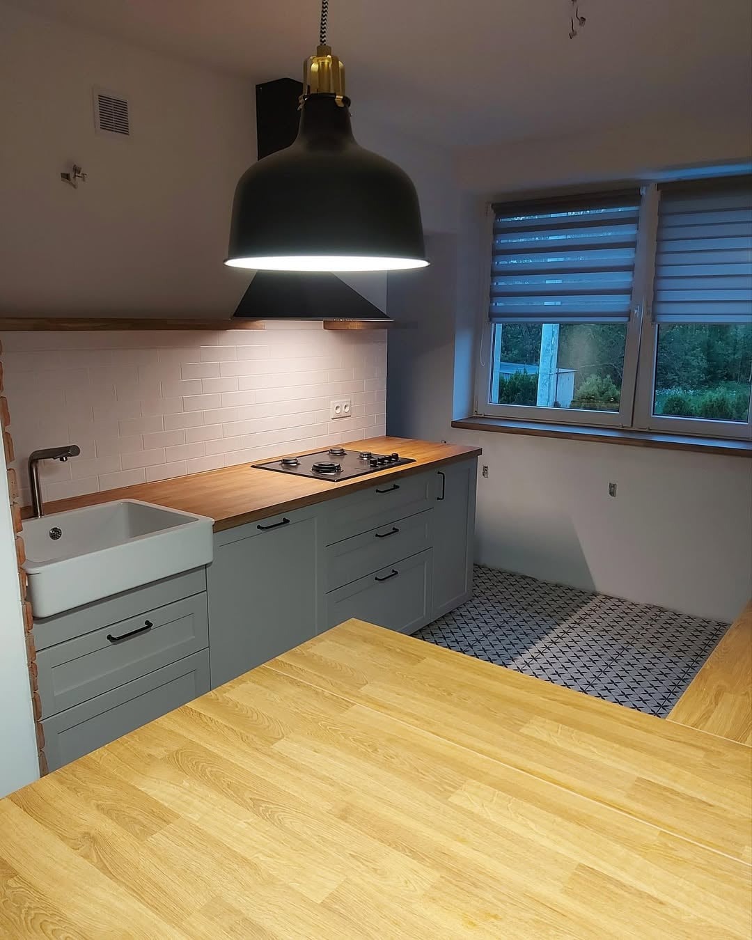 Jasna kuchnia po remoncie: szare szafki z drewnianym blatem, białe płytki na ścianie i wzorzysta podłoga. Nad blatem wisi duża, czarna lampa.