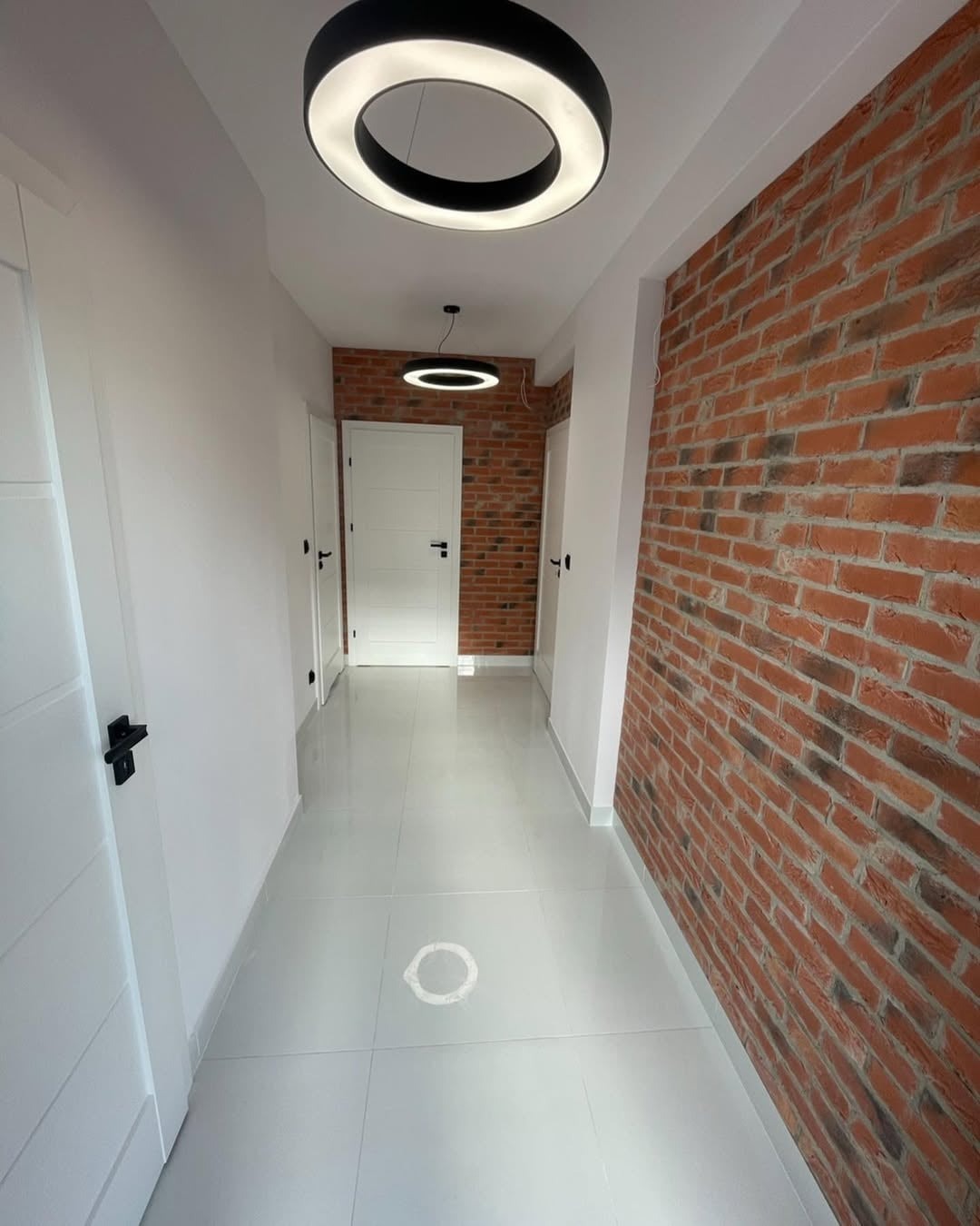Korytarz po remoncie: białe ściany, błyszcząca podłoga z płytek, ściana z cegły, okrągłe lampy sufitowe. Widok na drzwi i dalszą część korytarza.