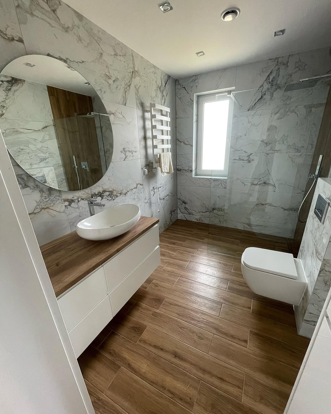Nowoczesna łazienka z marmurowymi płytkami, drewnopodobną podłogą i owalną umywalką nablatową. Widoczny grzejnik drabinkowy i kabina prysznicowa.