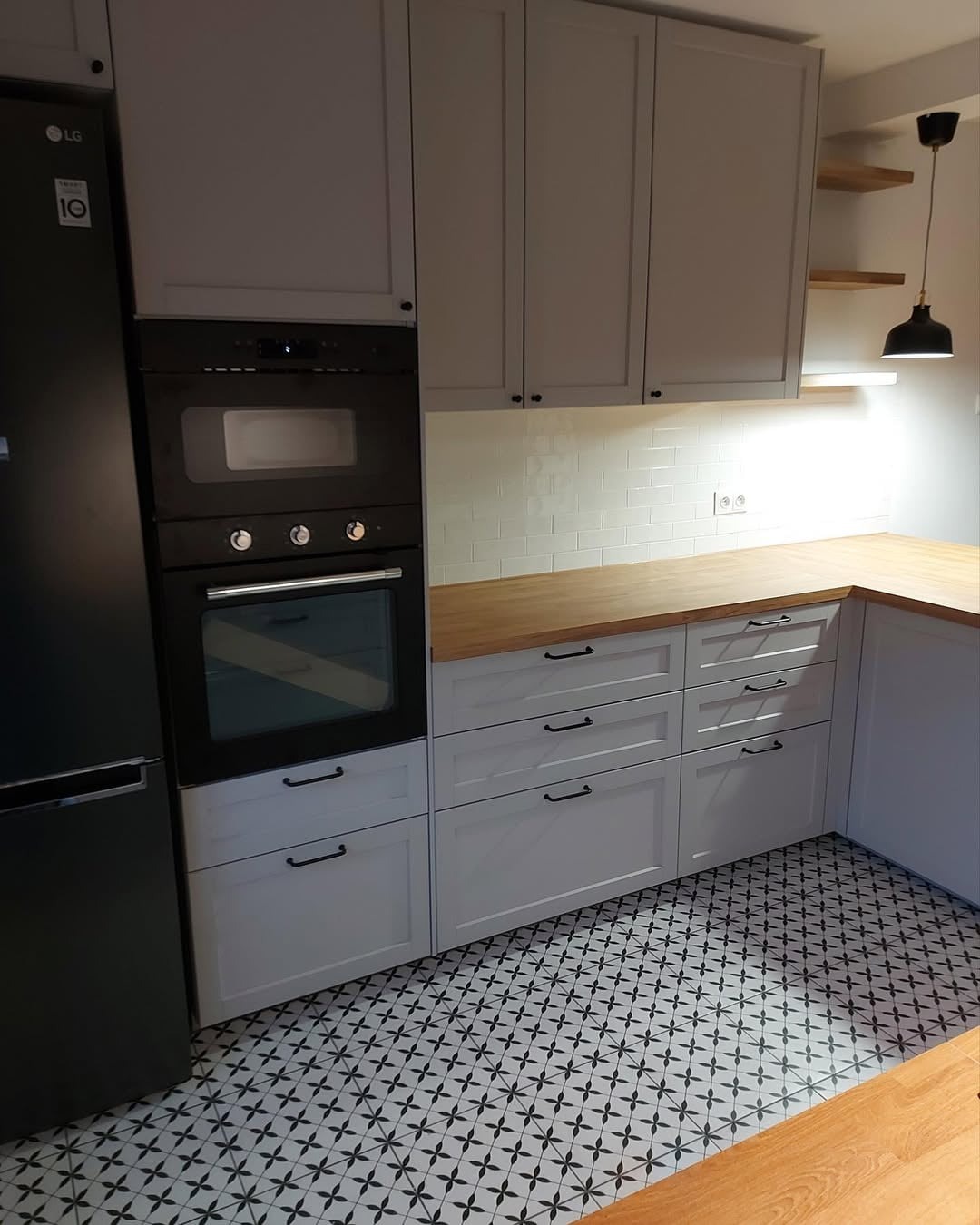 Jasna kuchnia z drewnianym blatem, czarnym piekarnikiem i lodówką LG. Wzorzysta podłoga i oświetlenie LED pod szafkami dodają nowoczesnego charakteru.