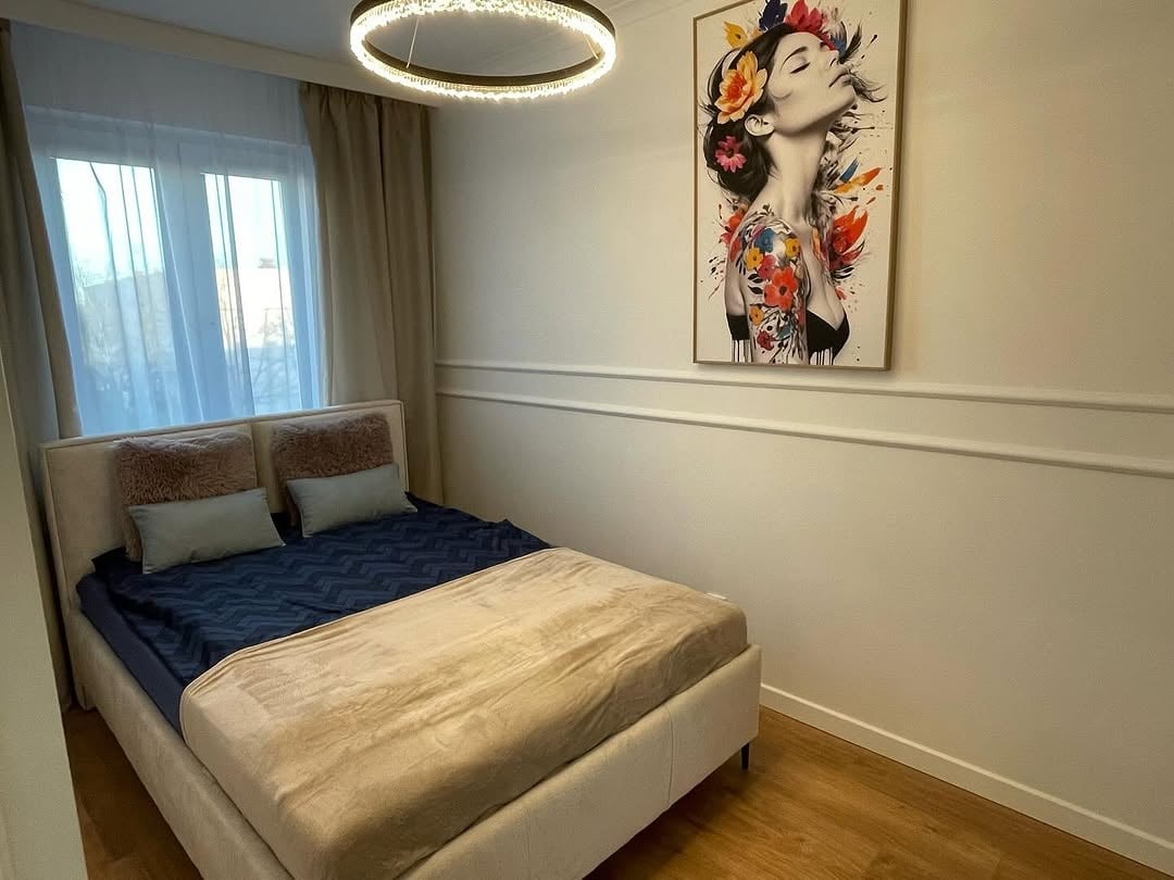 Jasna sypialnia z beżowym łóżkiem, dekoracyjną sztukaterią ścienną i obrazem kobiety z kwiatami. Okrągła lampa sufitowa dodaje nowoczesnego akcentu.