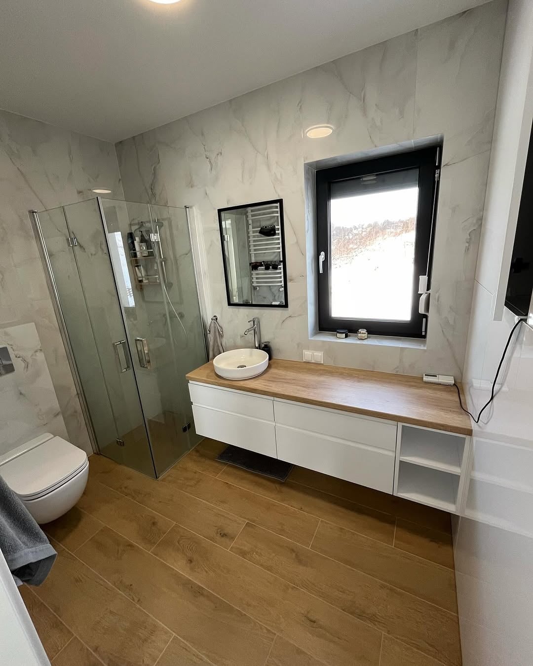 Nowoczesna łazienka z kabiną prysznicową, umywalką nablatową na szafce z drewnianym blatem i płytkami imitującymi marmur. Okno z czarną ramą.