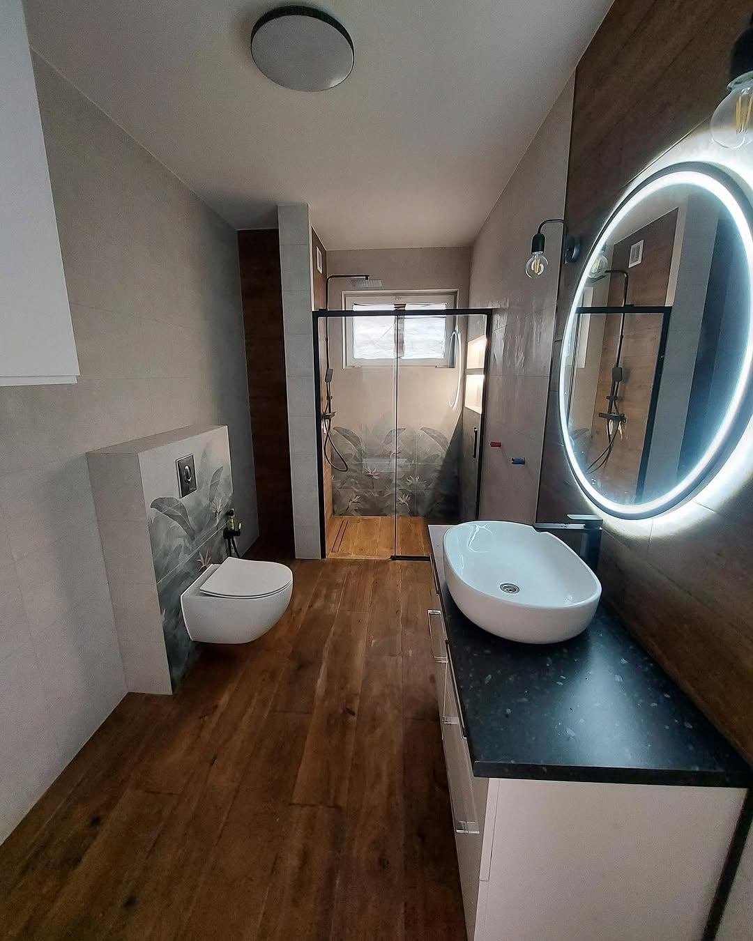 Nowoczesna łazienka z drewnianą podłogą i akcentami, kabina prysznicowa z motywem roślinnym, okrągłe lustro z podświetleniem LED i wisząca toaleta.