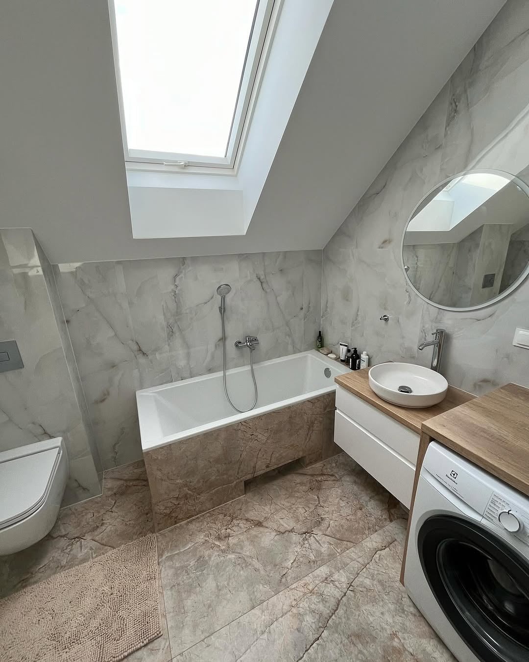 Jasna łazienka na poddaszu z oknem dachowym. Marmurowe płytki na ścianach i podłodze, wanna obudowana płytkami, umywalka nablatowa i pralka Electrolux.