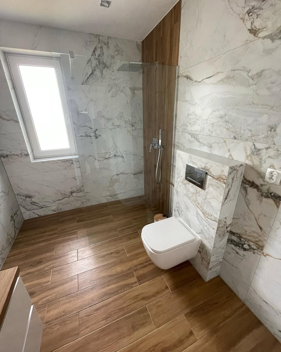 Nowoczesna łazienka z kabiną prysznicową typu walk-in, wykończona płytkami imitującymi marmur i drewno. Uwagę zwraca podwieszana toaleta i minimalistyczna armatura.