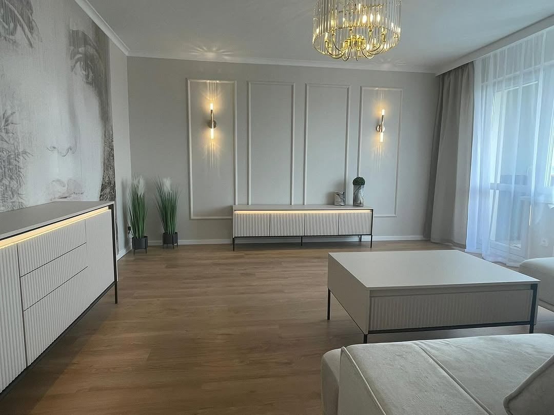 Elegancki salon z drewnianą podłogą, białymi meblami z ryflowanymi frontami, dekoracyjnym oświetleniem i abstrakcyjną grafiką na ścianie. Nowoczesny design.