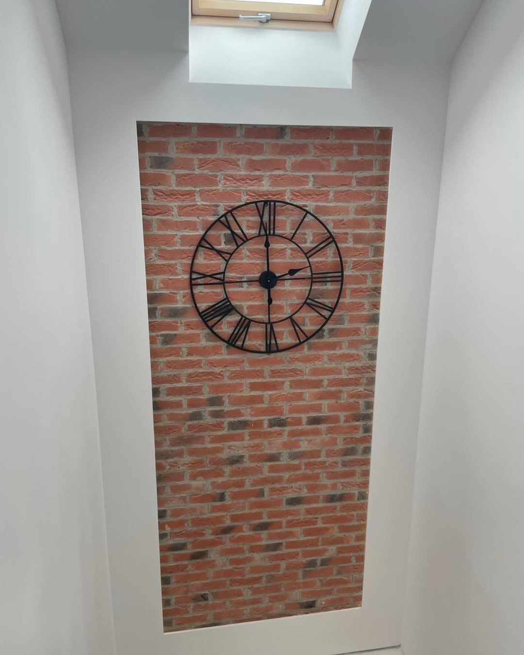 Wnęka z ceglaną tapetą i zegarem w stylu loft, oświetlona światłem z okna dachowego. Białe, gładkie ściany. Nietypowe ujęcie z dołu.