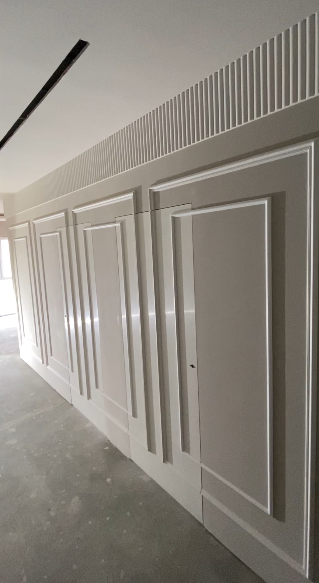 Eleganckie wykończenie ściany z panelami w stylu klasycznym, pomalowane na szaro z białymi listwami. Dekoracyjne pionowe żłobienia pod sufitem. Surowa podłoga.