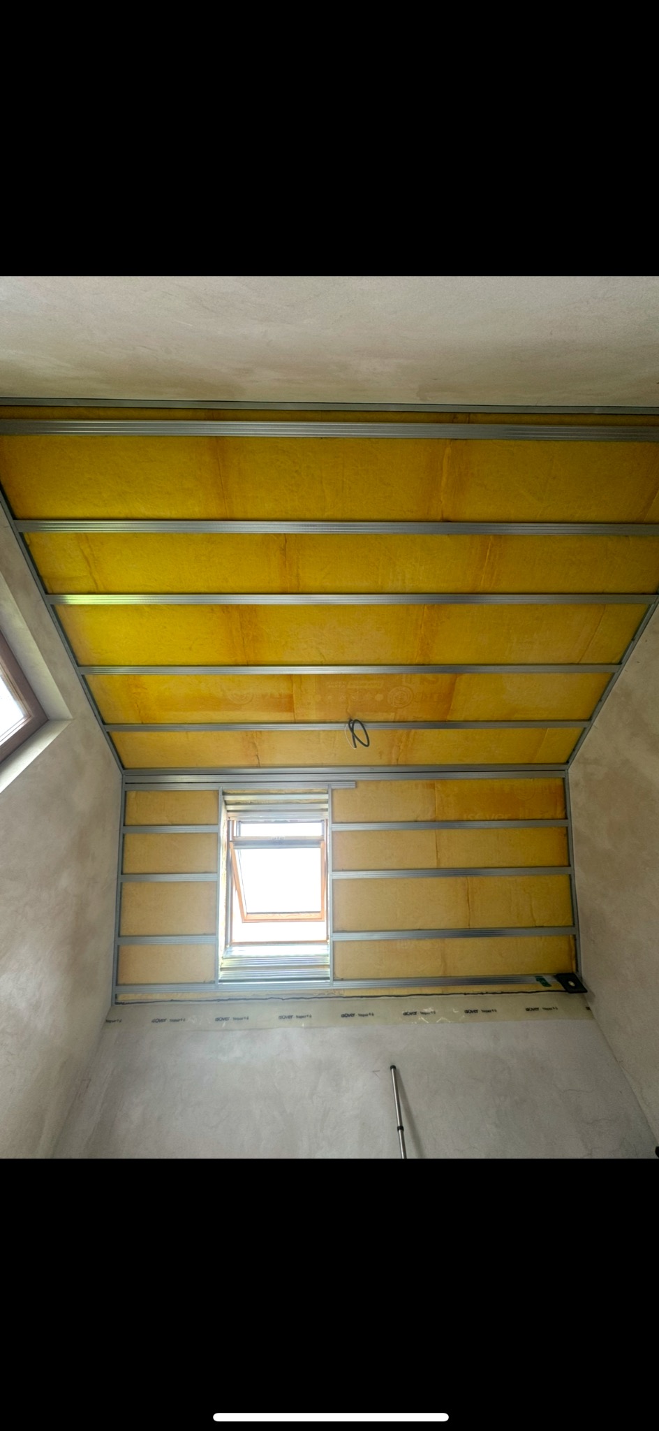 Montaż izolacji z wełny mineralnej na poddaszu z oknem dachowym, widoczne metalowe profile konstrukcyjne. Etap prac przed zakryciem płytami.