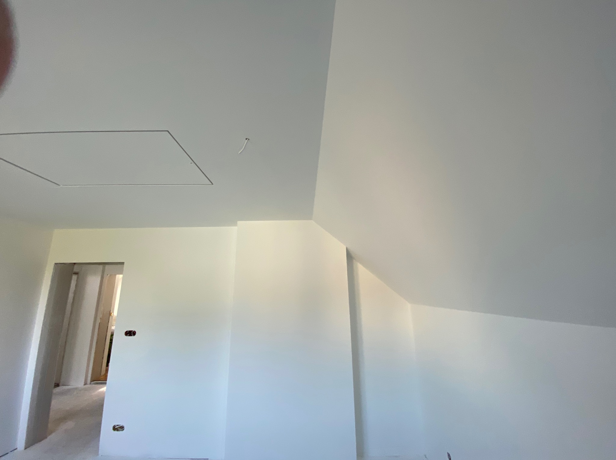 Białe, świeżo pomalowane wnętrze poddasza z widocznym włazem na suficie i otwartym przejściem do innego pomieszczenia. Gładkie ściany, gotowe do dalszych prac.