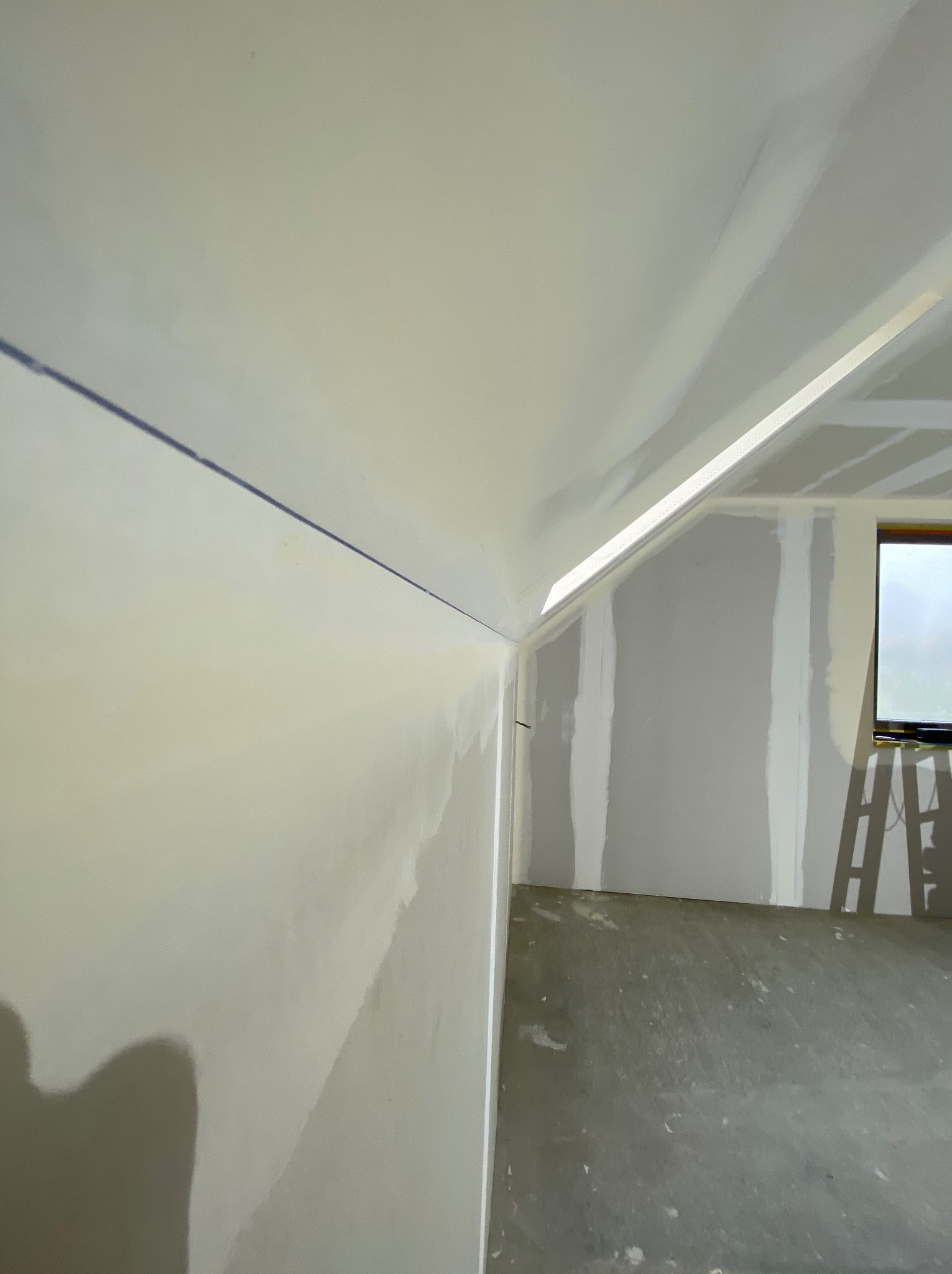 Pokój na poddaszu w trakcie prac wykończeniowych. Widoczne płyty karton-gips na ścianach i suficie, przygotowane do dalszej obróbki. W tle okno i drabina.