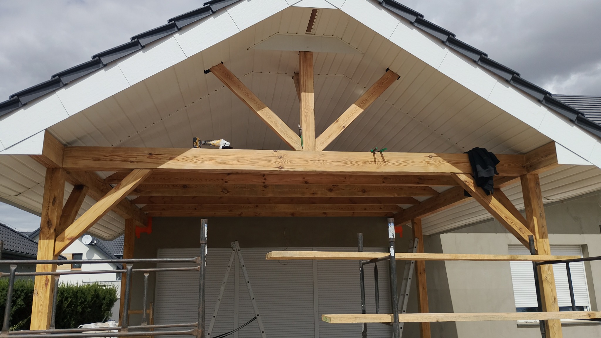 W trakcie budowy drewnianej konstrukcji z widoczną więźbą dachową i elementami obróbki blacharskiej. Na górze leży wkrętarka. Widoczne rusztowania i drabina.