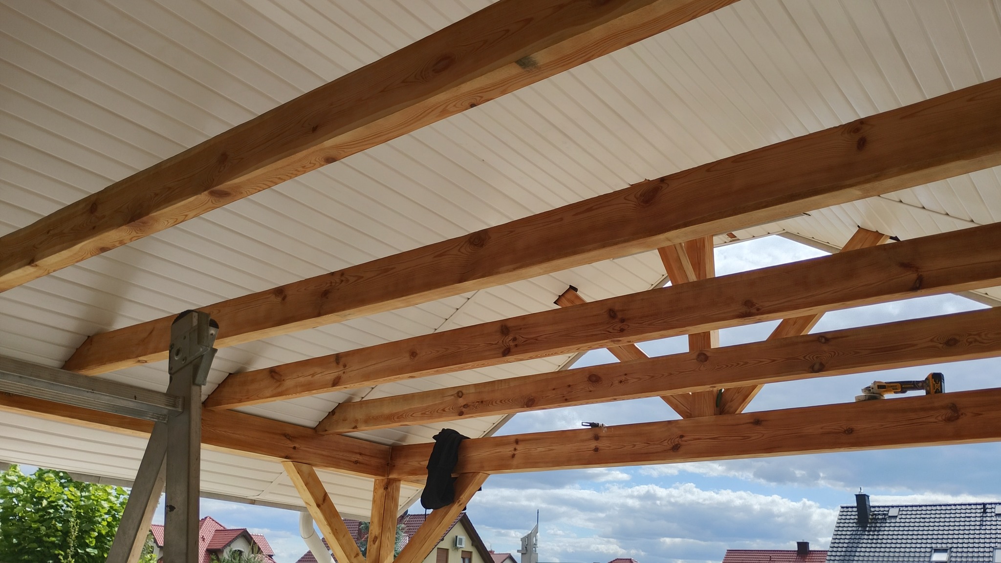 Drewniana więźba dachowa z białym podsufitką, widok z dołu. Na belce leży czarna bluza i wkrętarka. W tle domy i błękitne niebo z chmurami.