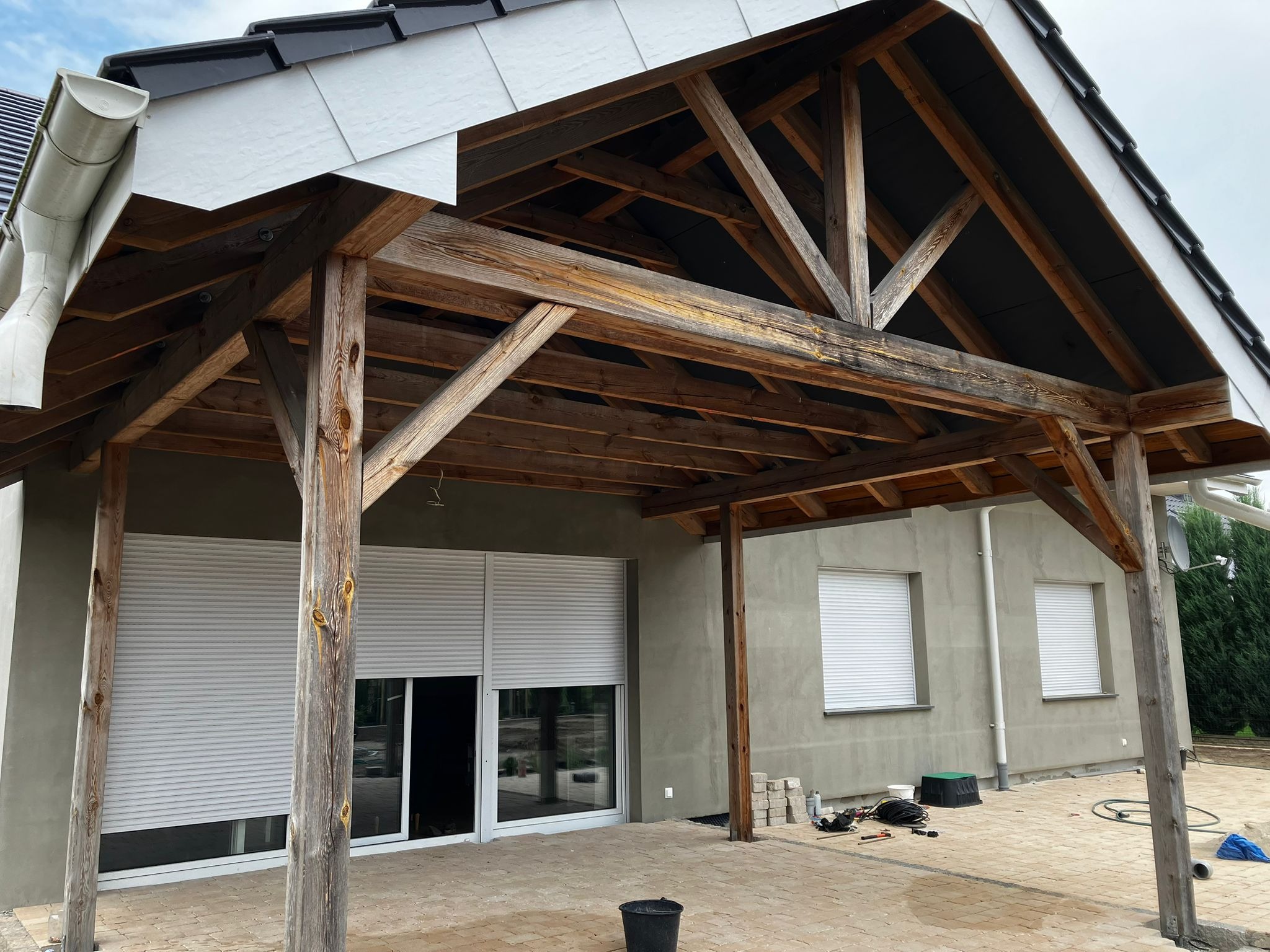 Drewniana konstrukcja zadaszenia tarasu w trakcie budowy, widoczne elementy więźby dachowej, rynna i fragment dachu z czarną dachówką. Częściowo zamknięte rolety okienne.