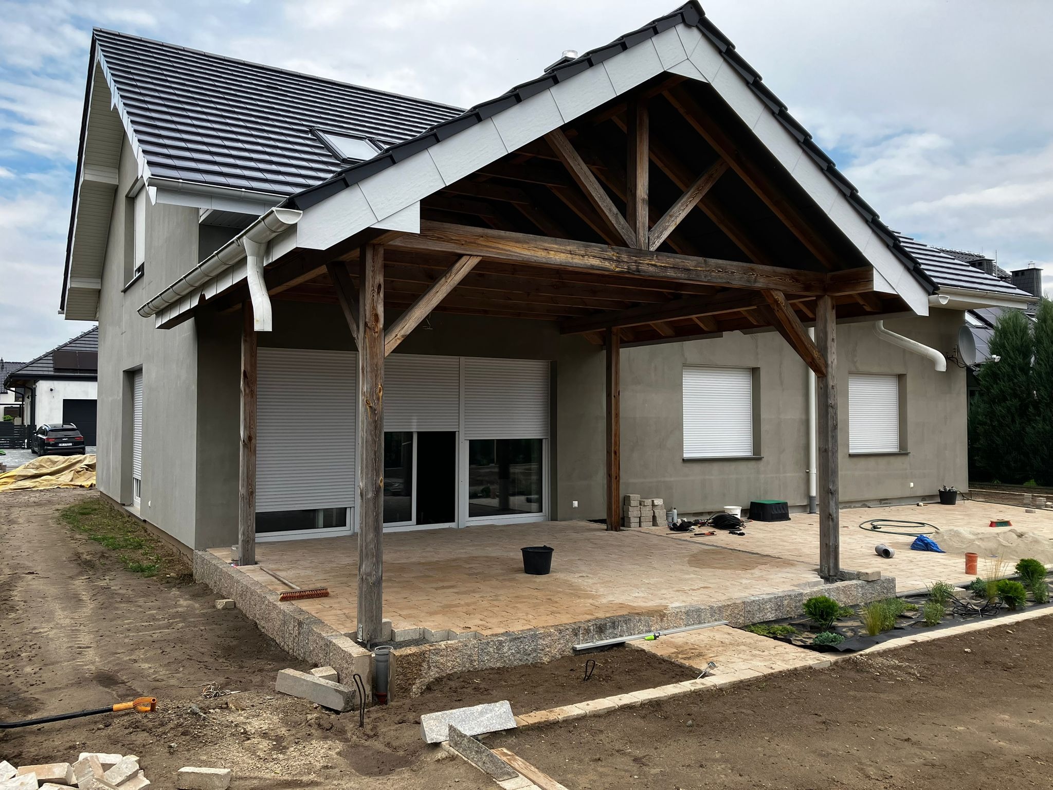 Nowoczesny dom z szarą elewacją i drewnianą pergolą w trakcie budowy tarasu z kostki brukowej. Widoczne rynny i dachówka.