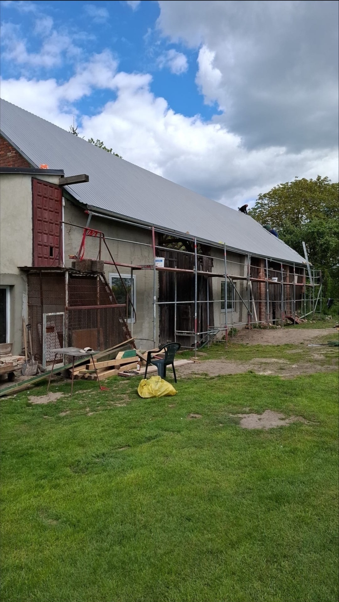 Budynek gospodarczy w trakcie remontu dachu, widoczne rusztowania i ekipa pracująca na dachu. Nowe poszycie w kolorze szarym. Prace w toku.
