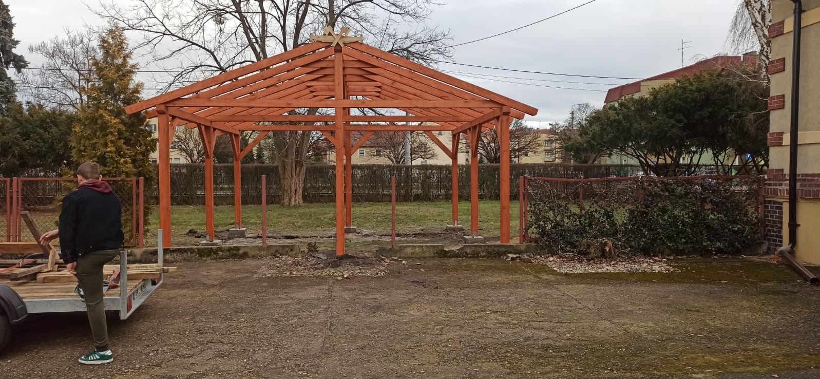 Drewniana konstrukcja altany ogrodowej w trakcie montażu, widoczne słupy i więźba dachowa, teren budowy z elementami narzędzi i przyczepką. Ujęcie w pochmurny dzień.