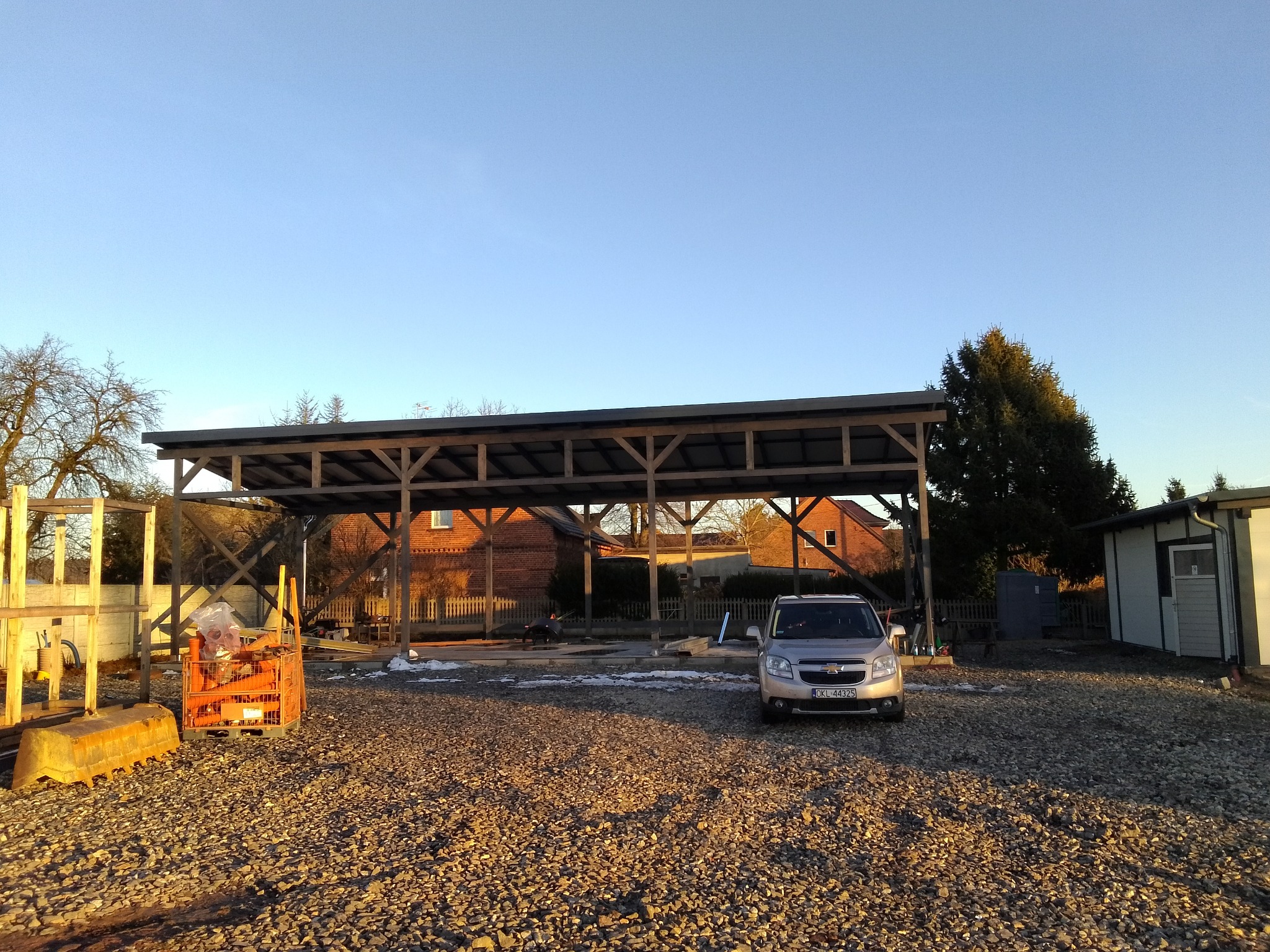 Drewniana konstrukcja zadaszenia na żwirowym podłożu, w tle budynki mieszkalne i samochód osobowy. Widoczne elementy konstrukcyjne i oświetlenie słoneczne.