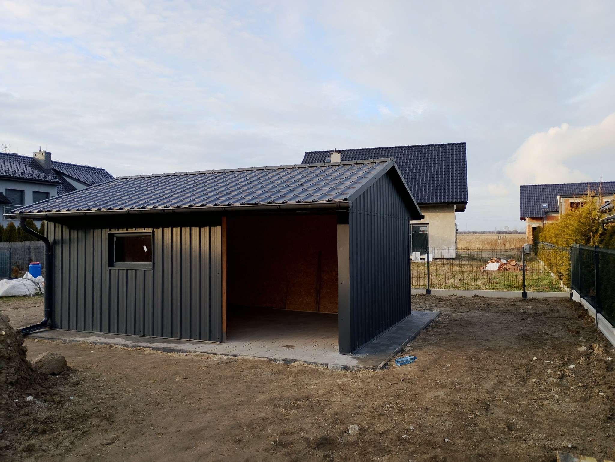 Nowoczesny, grafitowy garaż blaszany z dachem dwuspadowym i rynną, na tle domów jednorodzinnych w trakcie budowy, w pochmurny dzień.