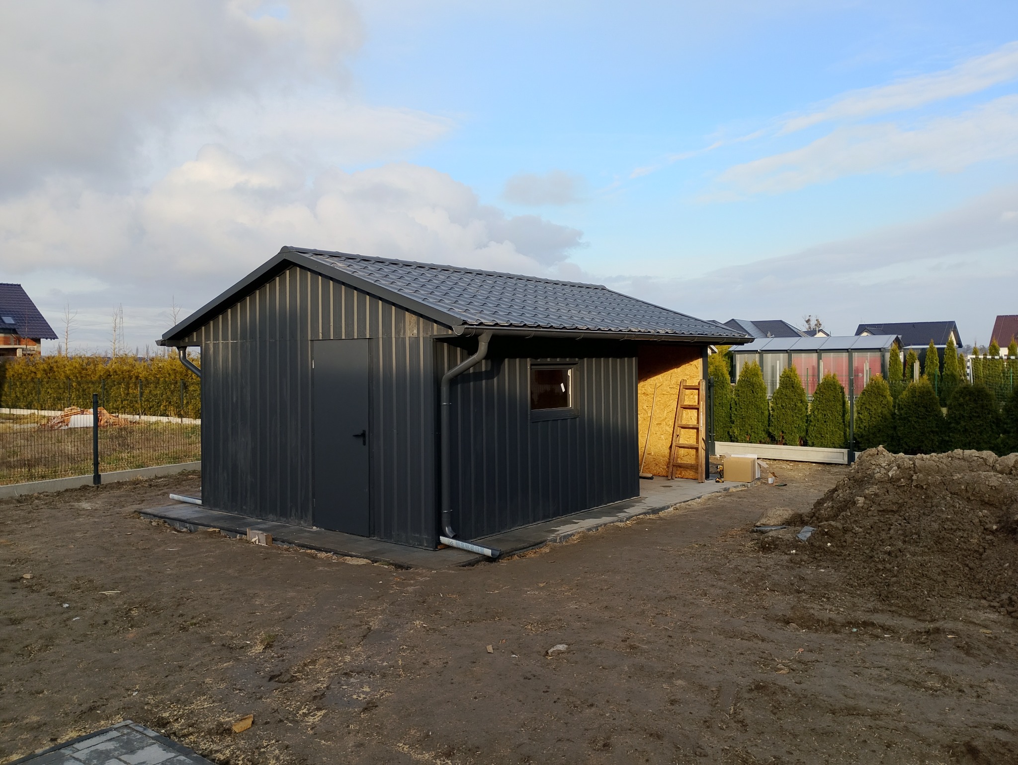 Nowoczesny, grafitowy budynek gospodarczy z blachy trapezowej i dachem z dachówki metalowej. Widoczne rynny, okno i drzwi. W tle domy i zieleń.