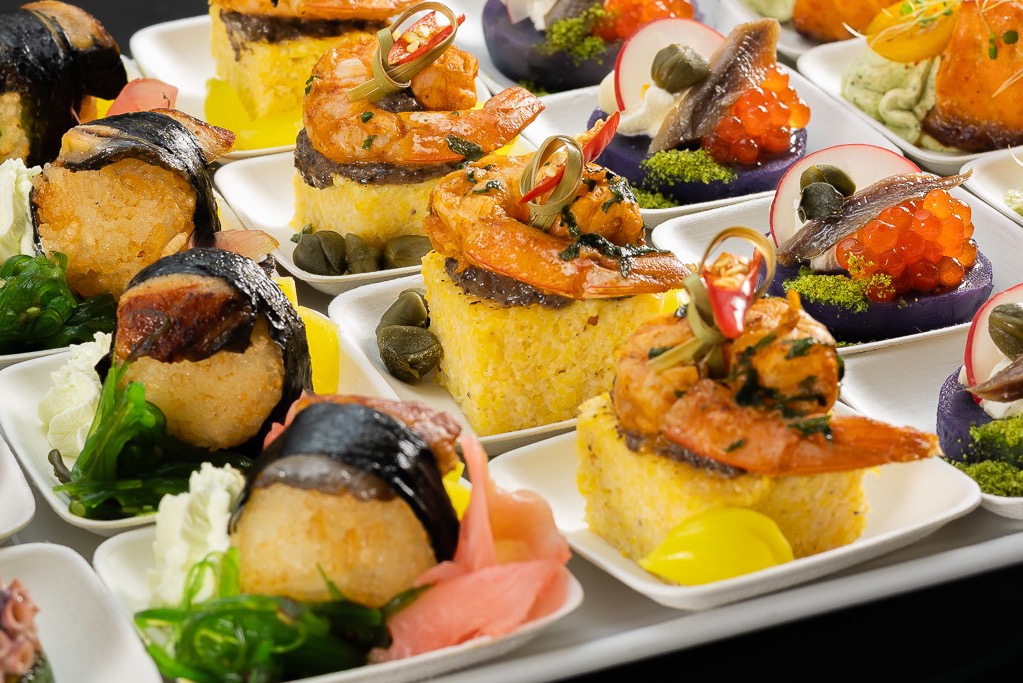 Ekskluzywne przekąski cateringowe na białych talerzykach: sushi z węgorzem, krewetki na polencie, fioletowe babeczki z kawiorem i anchois. Elegancki bufet.