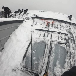 HuskyAlpine - Pracownik w uprzęży usuwa śnieg z dachu budynku z oknami dachowymi. Widoczne zaspy śnieżne i elementy systemu odwadniającego. Ujęcie z perspektywy pracownika.