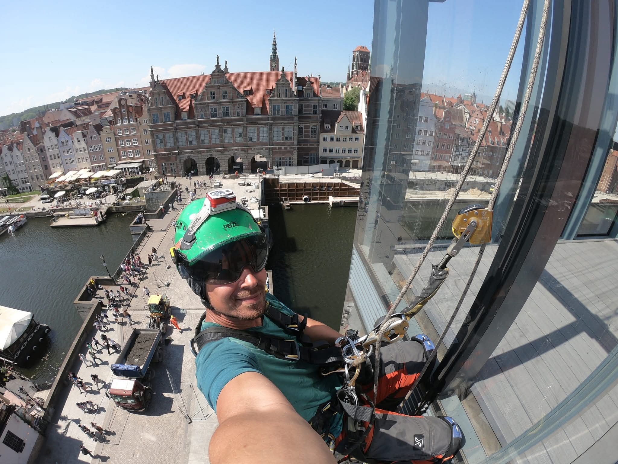 Pracownik w kasku i uprzęży, na tle panoramy miasta Gdańsk, wykonuje prace na wysokości na szklanej elewacji budynku. Widok z góry, ekspozycja na detale sprzętu.