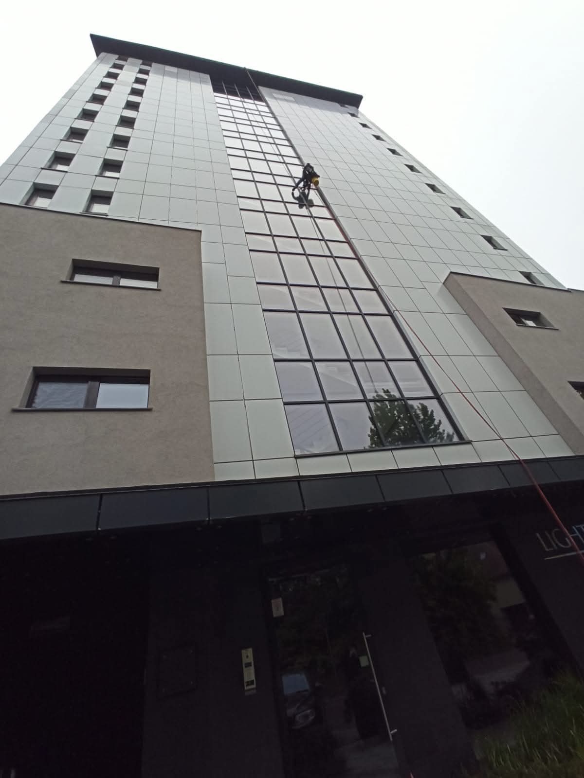 Pracownik na linach zabezpieczonych, czyści szklaną elewację wysokiego biurowca. Widok z dołu, pochmurne niebo w tle. Budynek z elementami z szarego tynku.