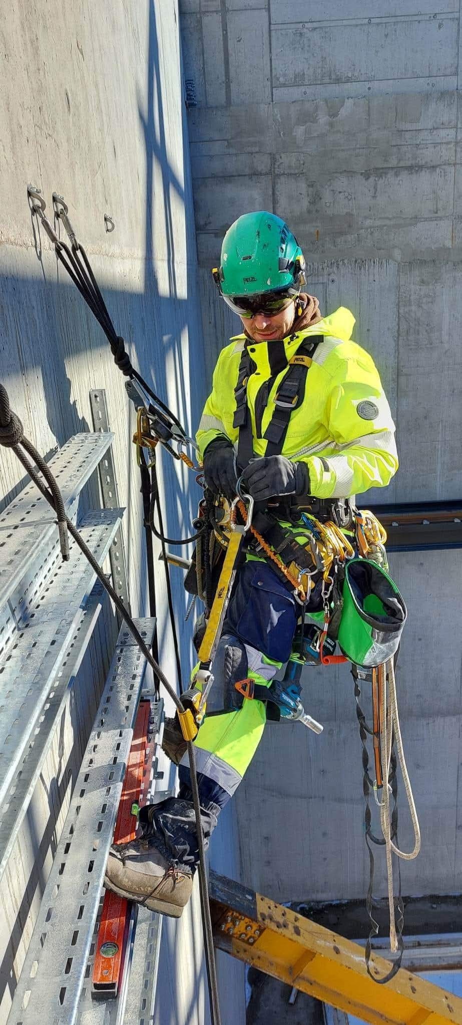 Pracownik w kasku i odzieży ochronnej, zabezpieczony linami, wykonuje prace na wysokości na tle betonowej elewacji. Widoczne narzędzia i elementy konstrukcyjne.