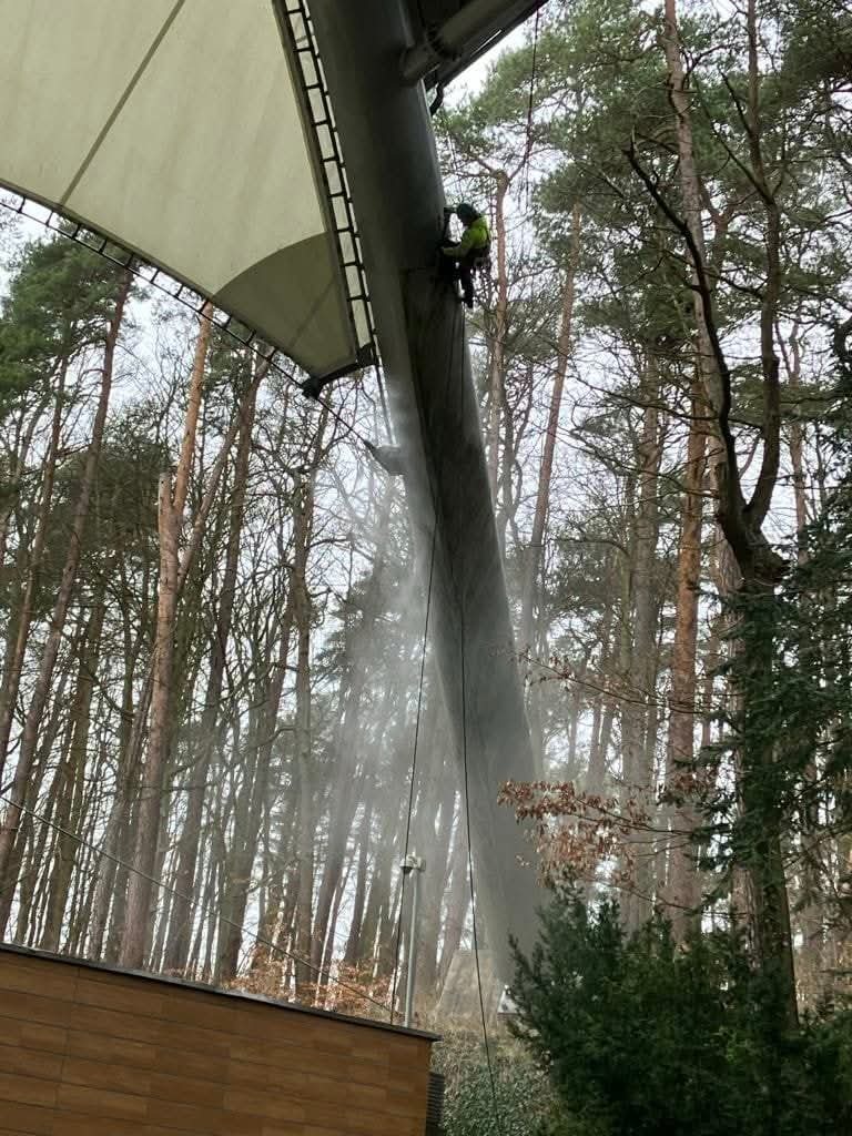 Pracownik na wysokości myje konstrukcję zadaszenia wodą pod ciśnieniem. Widoczne drzewa w tle i fragment budynku.