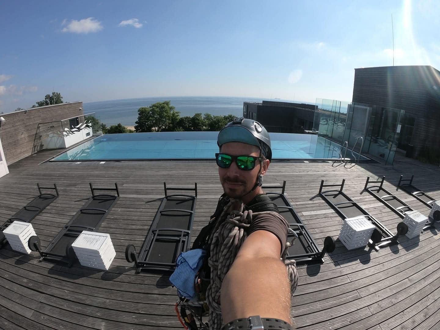 Pracownik w kasku i okularach, z linami, na dachu budynku z basenem i widokiem na morze. Ujęcie z dołu, szeroki kąt.