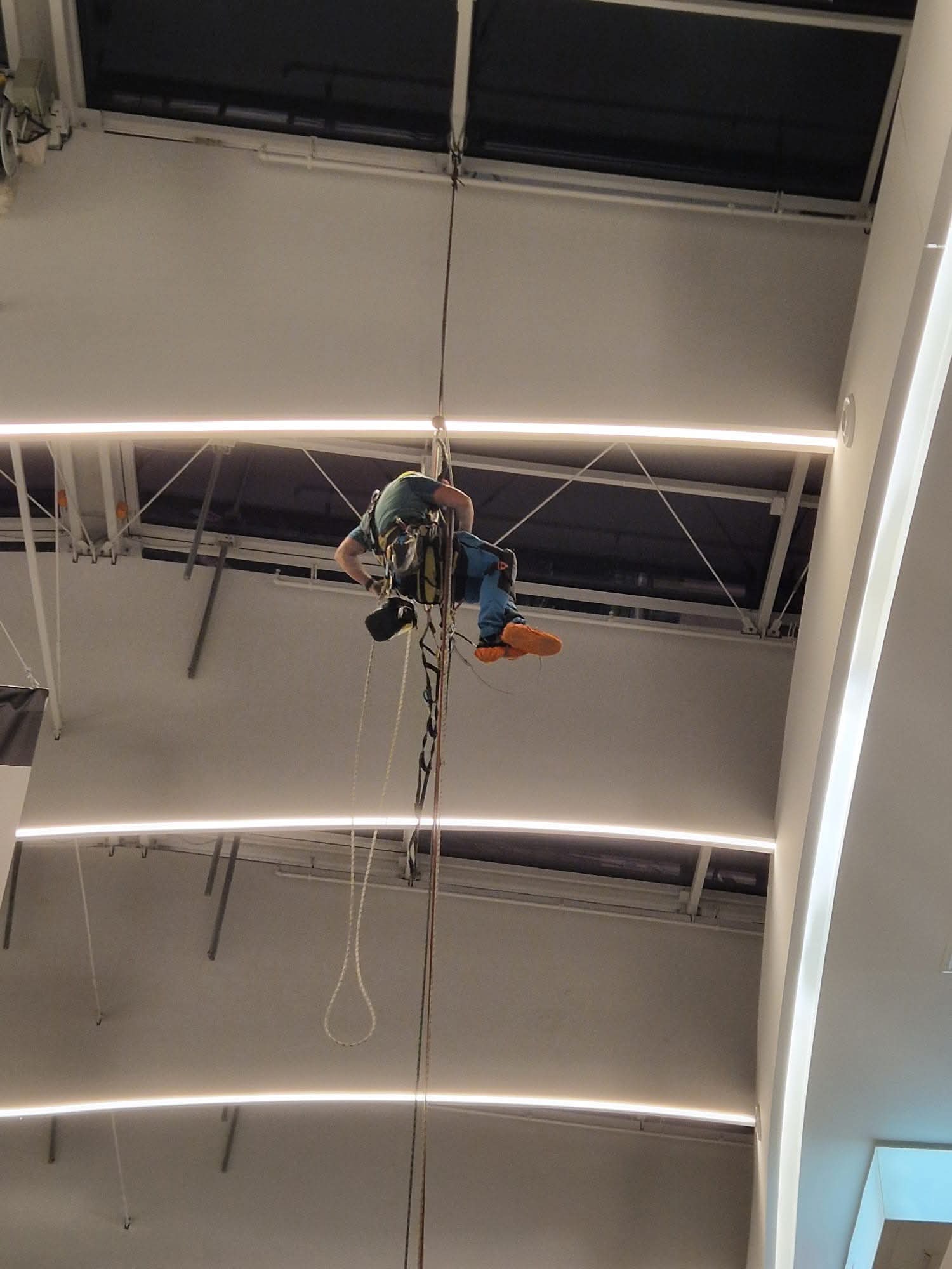 Pracownik na linach, w uprzęży, wykonuje zadanie na wysokości wewnątrz budynku. Widoczne oświetlenie sufitowe i elementy konstrukcyjne.