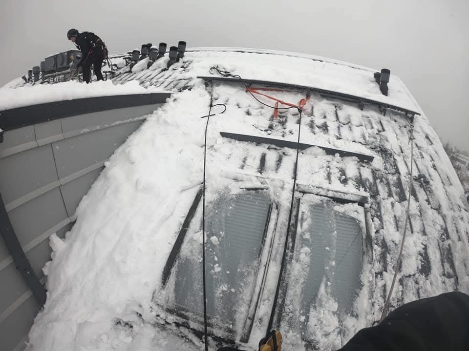 Pracownik w uprzęży usuwa śnieg z dachu budynku z oknami dachowymi. Widoczne zaspy śnieżne i elementy systemu odwadniającego. Ujęcie z perspektywy pracownika.