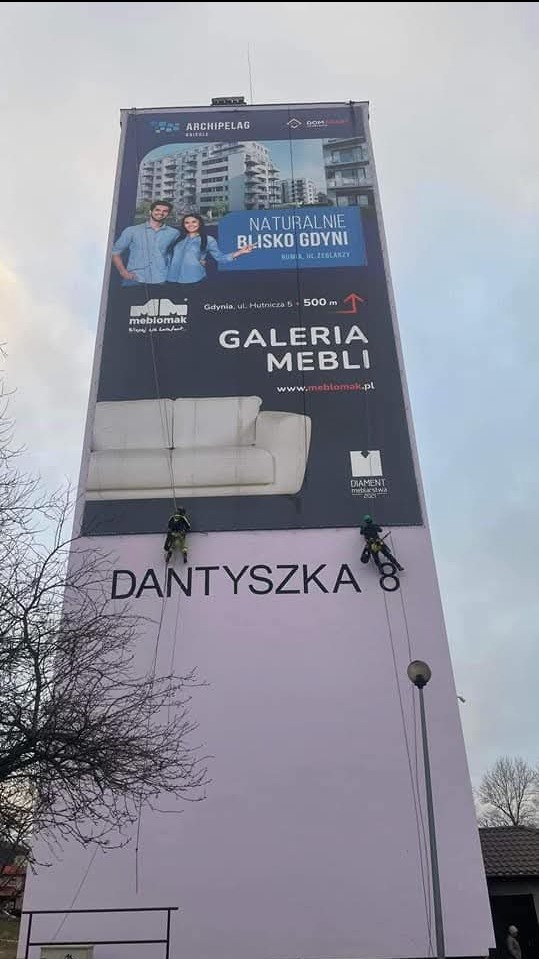 Dwóch alpinistów na linach podczas prac na wysokości, na tle ściany budynku z reklamą mebli 'Dantyszka'. Widoczne logo firm i hasło 'Naturalnie blisko Gdyni'.