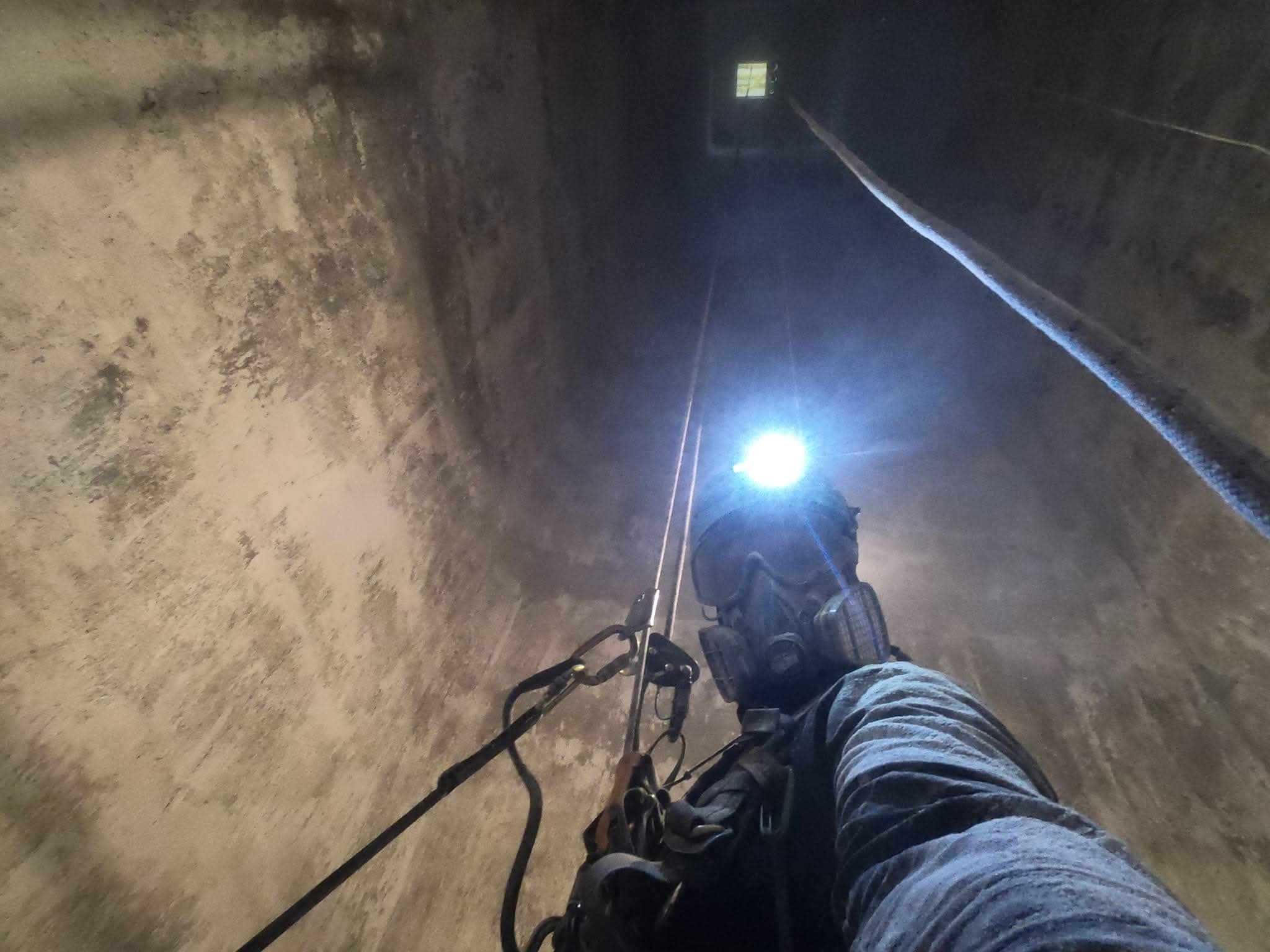 Pracownik w masce i uprzęży, oświetlony czołówką, w trakcie pracy na wysokości wewnątrz betonowej konstrukcji. Widok z dołu, liny i przewody w tle.