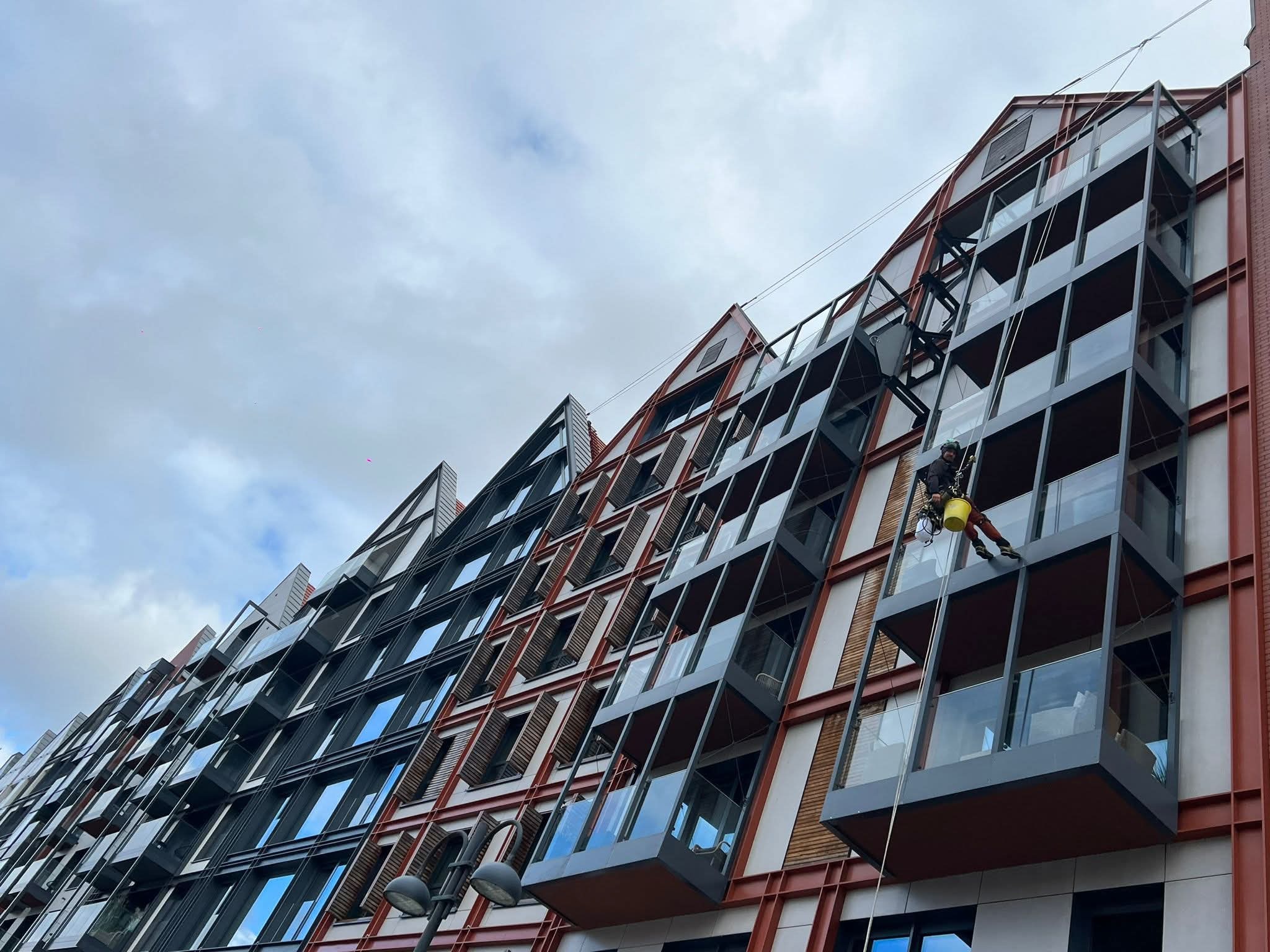 Pracownik na linach zabezpieczony uprzężą, wykonuje prace na wysokości na nowoczesnym budynku z przeszklonymi balkonami. Widok z dołu, pochmurne niebo.