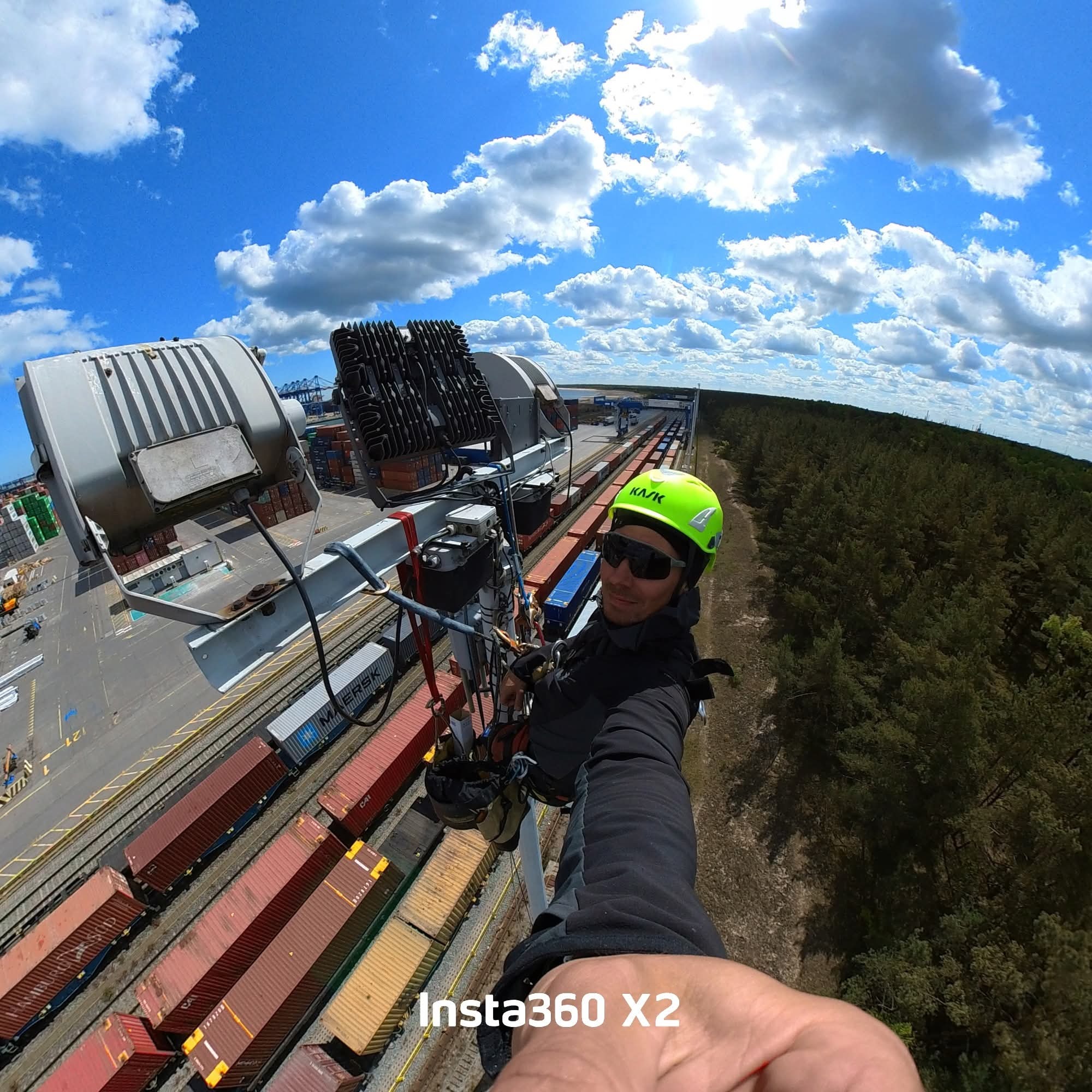 Mężczyzna w kasku i uprzęży na wysokości, prawdopodobnie podczas inspekcji oświetlenia przemysłowego na tle kontenerów i lasu. Szeroki kąt, słoneczny dzień.