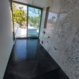 Marcin Głowiak - Wykończona łazienka z czarną marmurową podłogą i szarą marmurową ścianą z wnęką. Widoczne otwory na armaturę i duże okno z widokiem na zieleń.