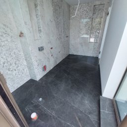 Marcin Głowiak - Wykończona łazienka z szarymi płytkami imitującymi marmur na ścianach i podłodze. Widoczne instalacje, wnęka w ścianie. Minimalistyczny design.